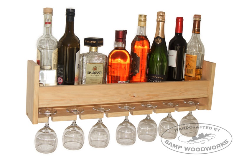 May include: A wooden wall-mounted wine rack with a shelf for bottles and a lower shelf for hanging wine glasses. The rack is made of natural wood and has a light brown finish. It is holding various bottles of wine and liquor, including a bottle of Disaronno.