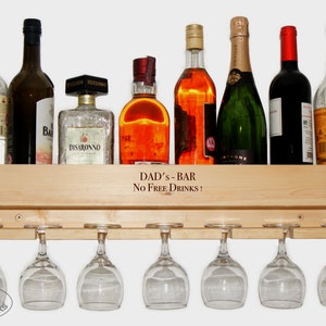 May include: A wooden wall-mounted wine rack with six glasses hanging underneath. The rack has a shelf for storing bottles and a sign that reads "DAD'S - BAR No FREE DRINKS!"