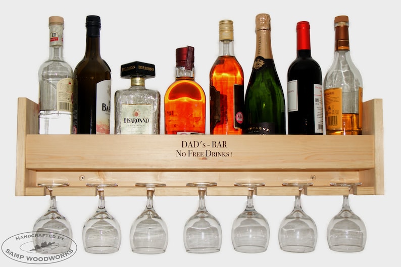 May include: A wooden wall-mounted wine rack with six glasses hanging underneath. The rack has a shelf for storing bottles and a sign that reads "DAD'S - BAR No FREE DRINKS!"