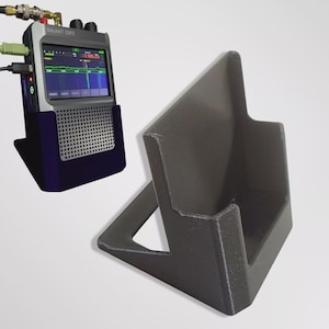 Puede incluir: Un receptor de radio Malahit DSP2 negro y gris con una pantalla que muestra un espectro de frecuencias, sostenido en un soporte azul oscuro. También se muestra un soporte gris oscuro separado, diseñado para sostener el receptor de radio.