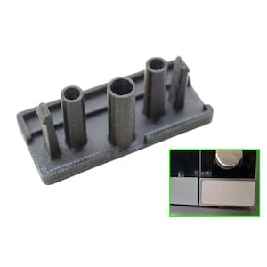 May include: A black plastic component with three cylindrical posts and a rectangular base. The component has a small rectangular tab on one end. The image also shows a close-up of a kitchen appliance.