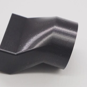 May include: A black, angled plastic connector with a cylindrical opening. The object has a textured surface and a geometric design, likely for connecting pipes or tubes. The item is set against a plain white background.