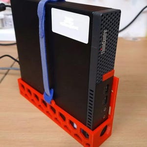 Peut inclure: Un ordinateur ThinkCentre noir est maintenu par une sangle bleue dans un support rouge imprimé en 3D. Le support présente un motif en nid d'abeille. L'ordinateur a un autocollant blanc et le logo ThinkCentre.