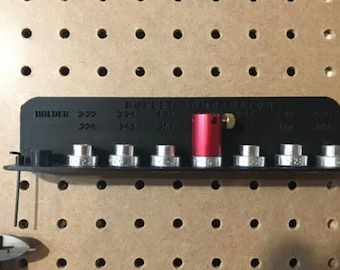 May include: A black "Bullet Comparator" tool with silver and red components. The tool has a series of silver holders and a red cylinder. Text on the tool indicates sizes such as .224, .243, and .257. The tool is mounted on a pegboard.