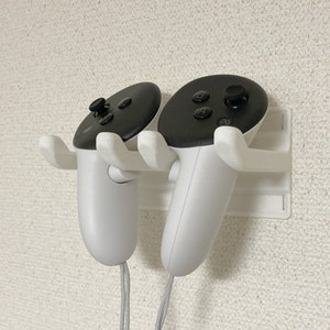 Puede incluir: Soporte blanco para pared para dos mandos de realidad virtual. Los mandos blancos tienen tapas negras con botones y un joystick. El soporte ofrece un almacenamiento ordenado.