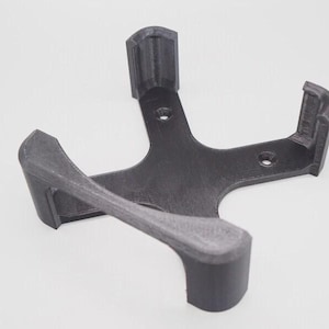 Puede incluir: Soporte de pared gris oscuro, impreso en 3D, para un dispositivo doméstico inteligente. El soporte tiene un diseño angular único con brazos curvos y una estructura de soporte central. Tiene orificios de montaje.