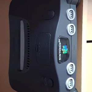 May include: A dark gray Nintendo 64 video game console. The console has a distinctive shape with rounded handles and several white buttons. The Nintendo 64 logo is visible on the front.