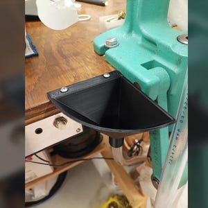 Pode incluir: Um funil preto triangular impresso em 3D, fixado a uma prensa de recarga. O funil é montado numa superfície de madeira com três parafusos. A prensa de recarga é de cor verde-azulado.