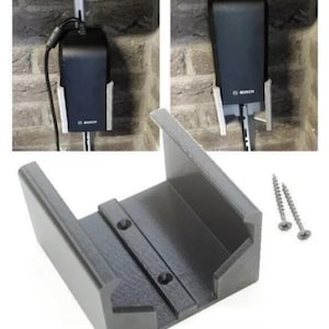 May include: A black Bosch power tool charger and mounting bracket. The charger is mounted on a wall with a white bracket. The bracket is gray and includes two screws.