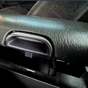 May include: Close-up of a black car interior armrest with a textured surface. The armrest features a recessed, rectangular compartment with a chrome-colored handle. The image highlights the details of the car's interior design.