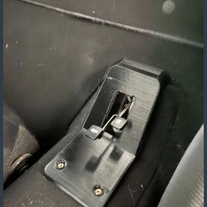 Puede incluir: Un mecanismo de ajuste de asiento de coche de metal negro con dos pequeños pedales y un resorte. El mecanismo está montado sobre una superficie negra y asegurado con tornillos. La imagen está tomada desde un ángulo bajo.