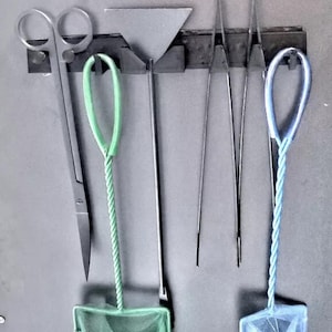 Könnte beinhalten: Aquarien-Wartungswerkzeuge, darunter eine Schere, ein Schaber, eine Pinzette und zwei Fischernetze. Die Werkzeuge sind an einem schwarzen Halter vor grauem Hintergrund montiert. Die Netze haben grüne und blaue Griffe.