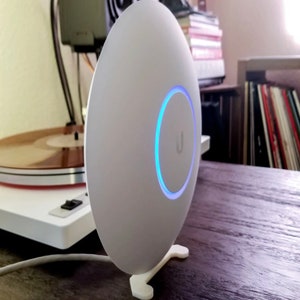 May include: A white, oval-shaped wireless access point with a blue illuminated ring and the letter "U" on the front. It is mounted on a white stand and sits on a wooden surface. The device is designed for network connectivity.