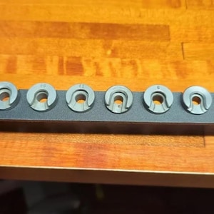 Puede incluir: Un conjunto de siete piezas metálicas grises, cada una con una forma curva y una abertura central, dispuestas sobre un soporte metálico negro. Las piezas están numeradas. El soporte está sobre una superficie de madera.