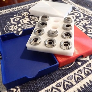 Puede incluir: Un conjunto de componentes metálicos plateados dispuestos en un inserto de espuma blanca. Los componentes tienen una forma única con una abertura central. Los contenedores son azules y rojos, sobre una tela estampada.