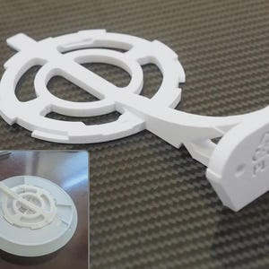 May include: A white, 3D-printed object with a circular design and a curved arm. The circular part has an open, geometric pattern. The arm leads to a rectangular base with two holes and a symbol. The object is on a textured surface.