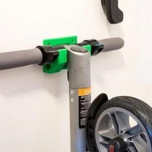 May include: A grey scooter handlebar with textured grips is mounted on a wall with a bright green bracket. The scooter's stem and a portion of the front wheel are also visible. The wheel has a black tyre.