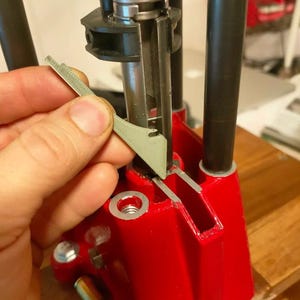 Può includere: Primo piano di una pressa di ricarica con uno strumento grigio che viene inserito. La pressa è principalmente rossa con componenti argentati e neri. Lo strumento è tenuto da una mano.
