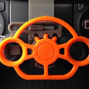 Op de afbeelding: Een oranje 3D-geprint stuurwielopzetstuk voor een gamecontroller. De opzet is ontworpen om over de knoppen en analoge sticks van de controller te passen, met een cirkelvormig ontwerp en een centrale naaf. De controller is zwart.