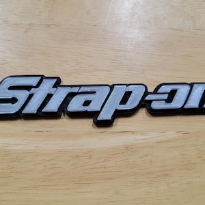 以下が含まれることがあります： 「Snap-on」の文字がスタイリッシュなフォントで表示された3Dプリントのサイン。文字は薄いグレーで黒い縁取りがあり、明るい木目調の背景に配置されています。このサインはおそらくロゴまたはエンブレムです。
