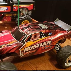 May include: A red and black RUSTLER VXL remote control car with large black tyres and a rear spoiler. The car has yellow and orange accents and is on a wooden surface. A black and red BLACK FOOT truck is in the background.