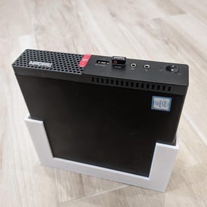 Könnte beinhalten: Ein schwarzer ThinkCentre Desktop-Computer mit rotem Akzent, gehalten in einem weißen Ständer. Der Computer hat eine perforierte Oberseite und verschiedene Anschlüsse. Ein Intel-Aufkleber ist auf der Vorderseite sichtbar.