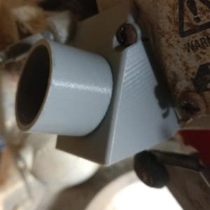 Pode incluir: Um componente cinza claro, impresso em 3D, com uma seção cilíndrica e um suporte em ângulo reto. O componente é montado em uma superfície de madeira, com um rótulo de aviso visível ao fundo. A seção cilíndrica tem um interior escuro.