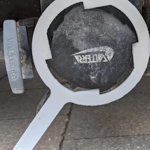 May include: A white plastic tool with a handle is used to open a black RV waste valve. The valve is labeled "Valterra" and the cap is labeled "Valterra". The tool is designed to fit around the cap for easy removal.