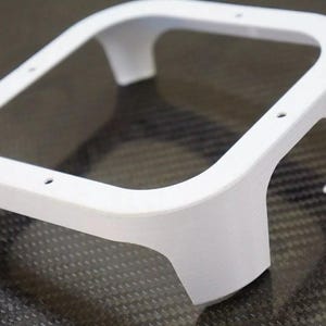 May include: A white, 3D-printed frame with a rectangular opening and curved sides. The frame has four legs and several small holes. The object is likely a component or housing for an electronic device or similar product.