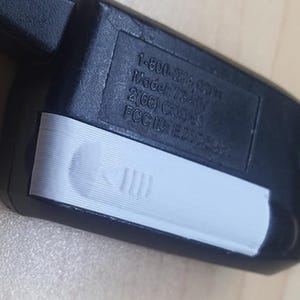 Puede incluir: Un mando a distancia negro con una sección blanca con un botón y una serie de líneas. El mando a distancia tiene un llavero y texto que incluye un número de teléfono, número de modelo e ID de la FCC.
