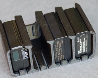 May include: A black battery holder with three black batteries. The holder is made of a dark gray material and has slots to hold the batteries securely. The batteries have text and a small screen.