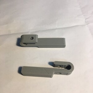 May include: Two gray, 3D-printed plastic clips. Each clip has a rectangular body with a curved end and a small hole. The clips are designed to hold something in place. The clips are on a white surface.