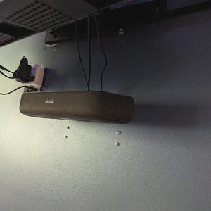 May include: A dark brown Roku soundbar mounted on a light blue wall. The soundbar is rectangular with rounded edges and the Roku logo is visible. Black wires hang down from the ceiling, connected to a power outlet.