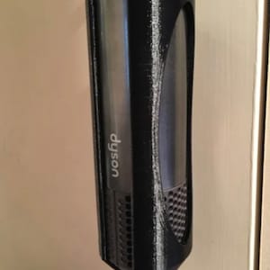 May include: A black 3D-printed hair dryer holder, designed to hold a Dyson hair dryer. The holder is mounted on a wall and features a curved design with the Dyson logo visible. The holder is made of black plastic.