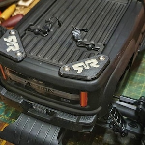 May include: A black remote-controlled truck with large, textured tires. The truck has the letters "SR" on the front corners and a ridged bed. The rear lights are orange. The truck is on a work surface.