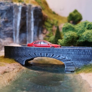 May include: A miniature red sedan crosses a grey stone bridge over a small stream. The bridge has an arched design and is surrounded by miniature trees and a waterfall in the background. The scene is a diorama.