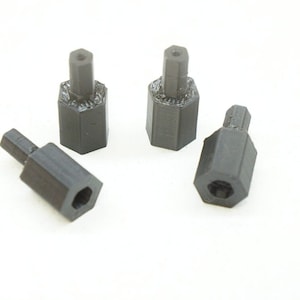 May include: Four black, hexagonal standoffs are arranged on a white background. Each standoff has a cylindrical post and a hexagonal base with a hole. The standoffs appear to be 3D-printed.