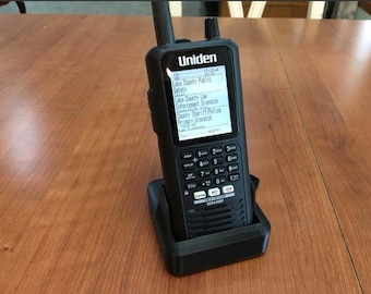 Pode incluir: Um scanner de rádio Uniden BC8436HP preto em um berço preto. A tela exibe texto, incluindo "Lake County Public Safety" e "County Sheriff/Police". O rádio tem um teclado e uma antena. O berço está em uma superfície de madeira.