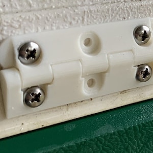 May include: A white plastic hinge with four silver screws. The hinge is mounted on a white surface, with a green surface visible below. The hinge is designed to allow movement between two surfaces.
