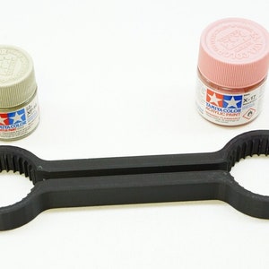 May include: A black 3D-printed tool for holding paint bottles, with two circular ends and a handle. Two small Tamiya Color acrylic paint bottles are present, one in a light green and the other in a pink color.