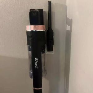 Puede incluir: Una aspiradora Shark negra y oro rosa con una boquilla para rincones adjunta. La aspiradora está montada en una pared. El logotipo de Shark es visible. La aspiradora tiene un botón de encendido.