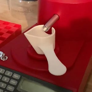 May include: A red reloading machine with a white scoop and a digital scale. The machine has a red hopper and a silver tube. A red tray is visible on the left side of the image. The scale has a digital display and several buttons.