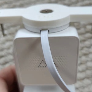 May include: A white security device with a square base and a circular top. The device has a white strap and two metal pins. The base has a series of small holes. The device is likely used for security purposes.
