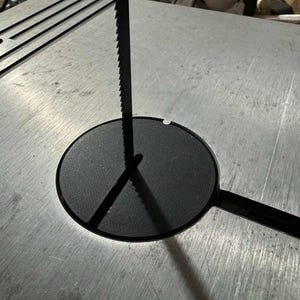 May include: A close-up of a band saw blade extending through a black insert on a metal table. The blade is black with sharp teeth. The table has a silver finish and a slotted section.