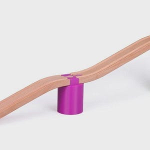 May include: Wooden toy train track with a curved design, supported by purple and green cylindrical blocks. The track is made of light brown wood and connects with purple connectors. A fun toy for children.