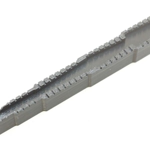 May include: A gray, 3D-printed tool with a rectangular channel and a series of small, evenly spaced teeth along the edges. The tool has a tapered design, with one end wider than the other. The object is on a white background.