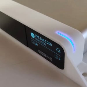 May include: A white and silver electronic device with a small black screen displaying network information. The screen shows an IP address and other data. A blue light is illuminated on the top edge of the device. The device has a modern, minimalist design.