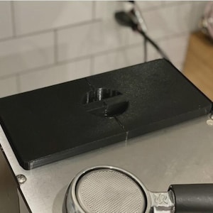 May include: A black, rectangular espresso machine accessory with a recessed area. The accessory sits atop a stainless steel surface, with a portafilter visible below. The background includes a white tiled wall and a power cord.