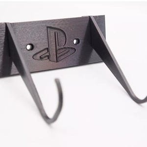 May include: A black wall-mounted game controller holder. The holder features the PlayStation logo and two curved arms to hold the controller. The holder is made of a matte black material and has two holes for mounting.