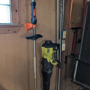 Könnte beinhalten: Ein orangefarbener und schwarzer Echo-Trimmer und ein gelb-schwarzes Ryobi-Laubgebläse sind an einer Holzwand befestigt. Ein silberfarbener Trimmer befindet sich links. Weitere Werkzeuge sind sichtbar.